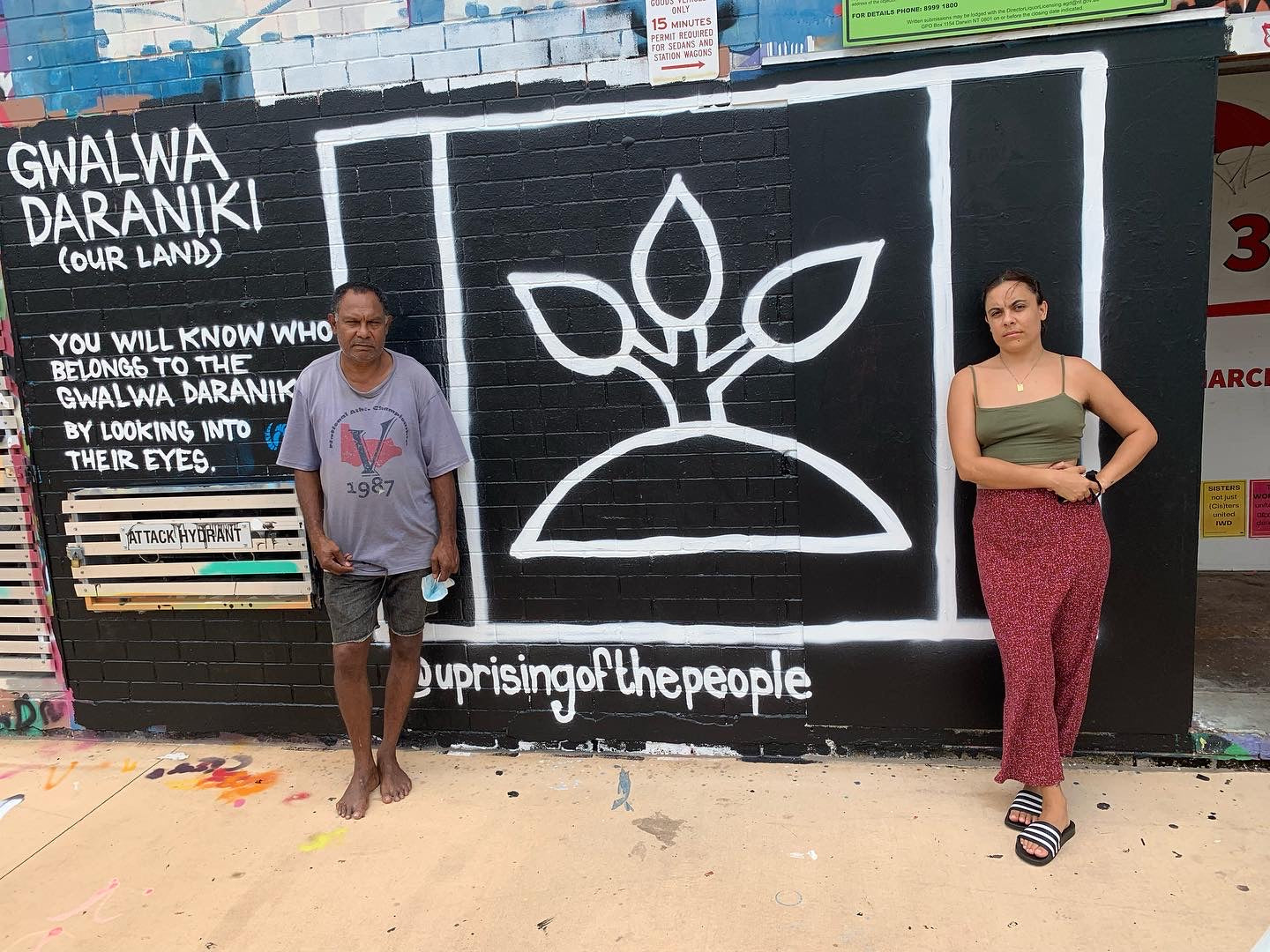 Photo of Father and Daughter standing in front of street art that reads "our land: you will know who belongs to our land by looking into their eyes". Next to the text is an image of the Larrakia flag. 