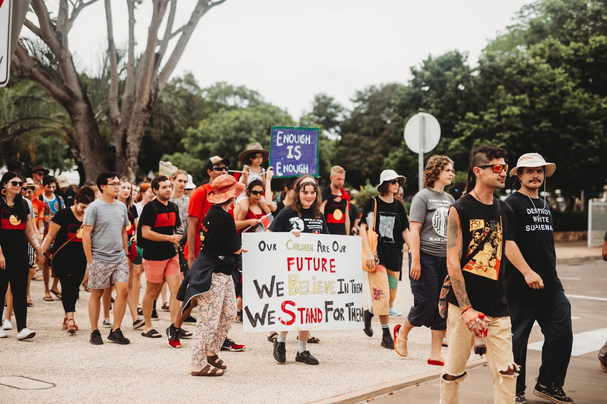 A photo from the Darwin BLM protest. A crowd of people walking on the road. A man wears a shirt that reads "destroy white supremacy". A sign reads "enough is enough". Some people are smiling other are serious.