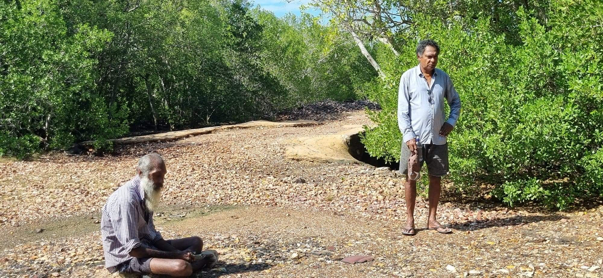 Two old men; one sitting crossed legged on the ground, the other standing. They are on a track that is leading through the mangroves.