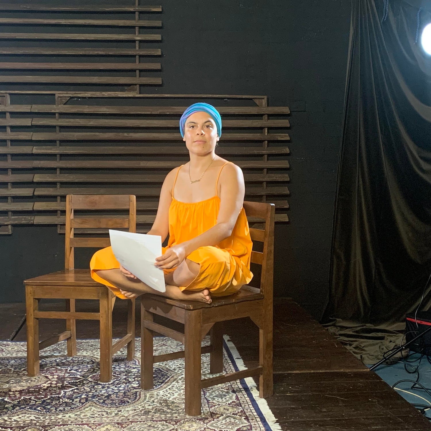 Image of young woman (Mililma May) sitting crossed legged on a chair. she wears a long orange dress, blue head scarf and holds a piece of paper in both hands. she stares past the camera with slight smile.