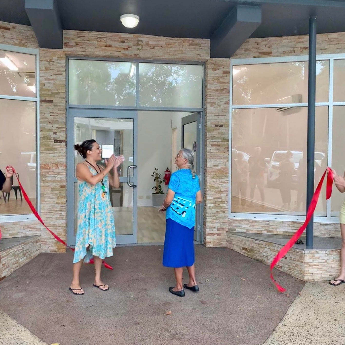 Mililma and Nana Mary smiling and clapping. They are standing in front their community center. Red ribbon is mid fall after being cut. 