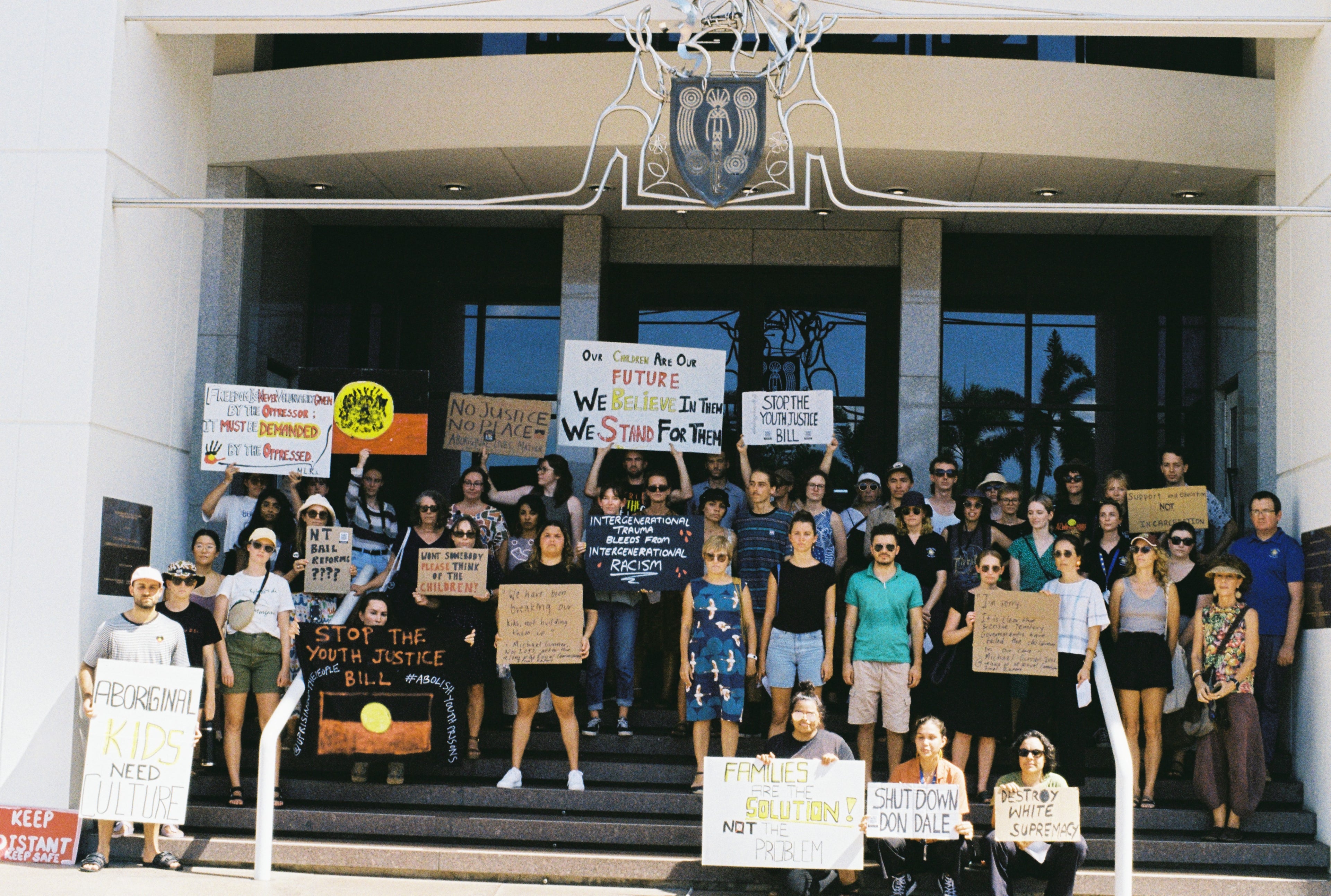 Image of a crowd on people on the steps of NT Parliament House. They are holding signs of the Aboriginal flag and with words like Shut Down Don Dale. the people have serious faces.
